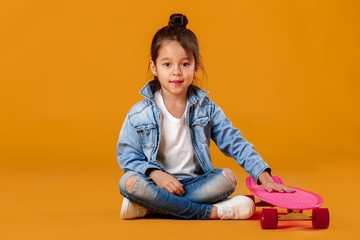 Naklejka premium Stylish little child girl with skateboard in jeans clothes on orange background