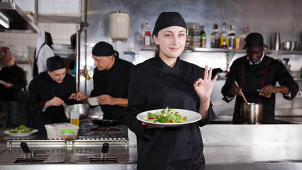 Positive female cook with cooked dish in restaurant kitchen