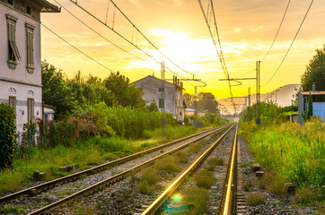 Fototapeta na wymiar Railway tracks with old buildings on sides, wires above and Tuscany hills and mountains with dramatic cloudy yellow orange sky background, evening, dusk, twilight view, near Lucca town, Italy