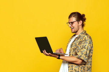 Portrait of a smiling handsome male in eyeglasses using laptop computer isolated over orange...