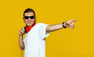Cheerful man with headphones standing on yellow background and pointing away with index finger.
