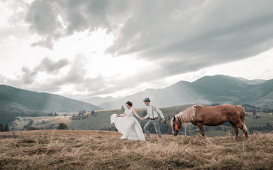 Fototapeta na wymiar Rural wedding in the mountains. newlyweds dressed in wedding clothes with a horse on the background of mountains. Mountain range at sunset. Beautiful couple with mountains amazing view