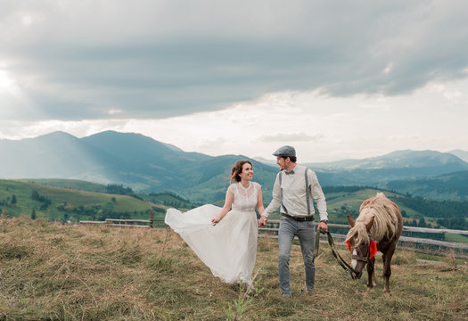 Beautiful bride and groom look at each other holding horse on the hill of mountains rocks on beautiful landscape