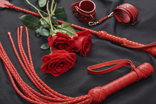 Playing BDSM games. Top view of bdsm leather kit (handcuffs, whip) and roses against of black silk fabric