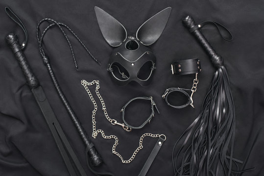 Explore the world of kinky play. Top view of bdsm leather kit (whips, handcuffs, mask and chain) against of a black silk.