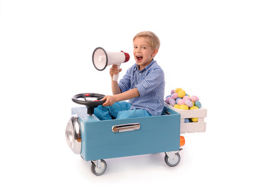 Child and Easter. A young blond boy rides a small blue toy car and carries a box of colorful eggs, holding a megaphone in his hand. Waist up portrait, facing camera. Studio photos.