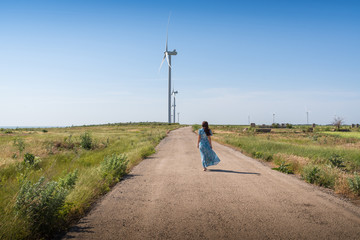 Beautiful woman with long hair in a blue dress walks on road between green fields and wind turbines on blue sky background