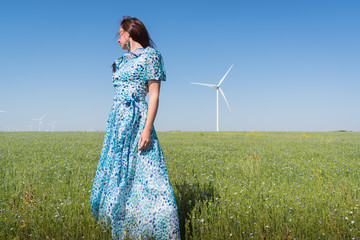 Beautiful woman in blue dress on green field with wind generator turbines and blue sky background
