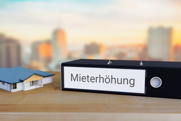 Miete erhöhen. Akten-Ordner beschriftet mit dem Wort Mieterhöhung liegt neben einem Haus-Modell auf einem Schreibtisch. Skyline einer Stadt im Hintergrund.