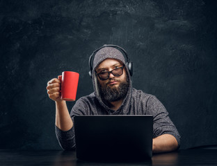 A hacker sits behind a laptop and holding a cup of coffee in a dark office and stares intently at...