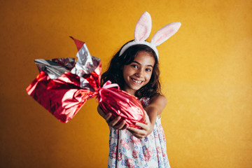 Cute little child wearing bunny ears on Easter day on color background. Girl holding chocolate...