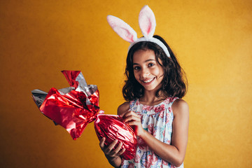 Cute little child wearing bunny ears on Easter day on color background. Girl holding chocolate...