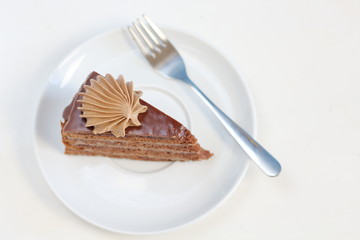 A piece of delicious chocolate cake with chocolate cream decoration on a white plate with dessert fork top view. Menu style