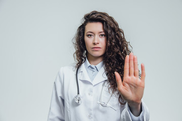 A young beautiful woman doctor gesturing stops. Probably a female doctor shows a gesture stop over a gray background.