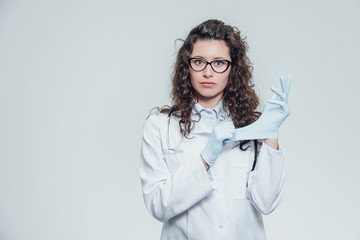 Young woman doctor wearing glasses in blue gloves looks intently into the camera. Isolated on a gray background