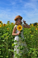 Mädchen mit Sonnenblume steht im Feld voller Blumen und macht lustiges Gesicht und Handbewegung