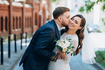 Handsome groom and beautiful bride sincerely posing on the streets of the old city. Groom kiss happy bride
