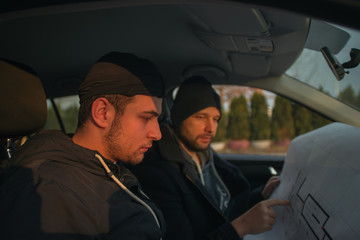 Two robbers sitting inside a car with guns looking at the blueprint of the building they are about...