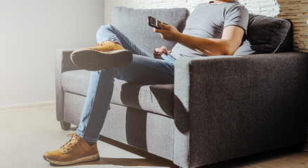 The man sits on a gray couch and switches programs on the TV. The concept of rest and relaxation after work. The man spends his free time watching TV, searching for a movie, killing boredom.