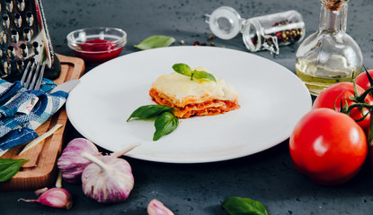 Lasagne on a white plate. On the stone table lies lasagna, spices and food ingredients. The concept of eating and preparing lasagna in tomato sauce.