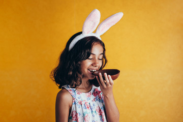 Cute little child wearing bunny ears on Easter day on color background. Girl eating chocolate...