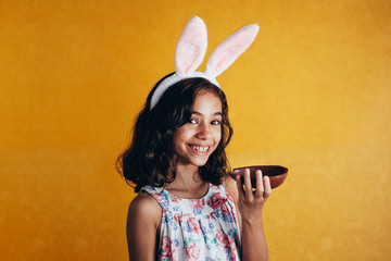 Cute little child wearing bunny ears on Easter day on color background. Girl eating chocolate easter egg