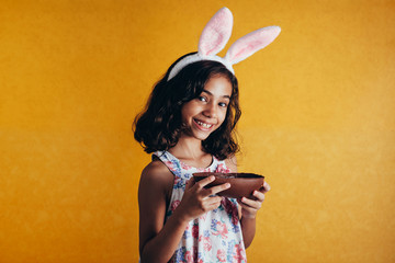 Cute little child wearing bunny ears on Easter day on color background. Girl eating chocolate easter egg