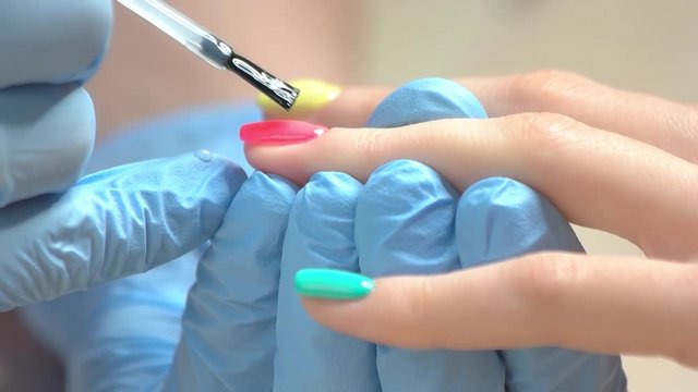 Slow motion nail covering with varnish. Close up cosmetician applying transparent fixer to client nail in beauty salon. Woman receiving manicure.