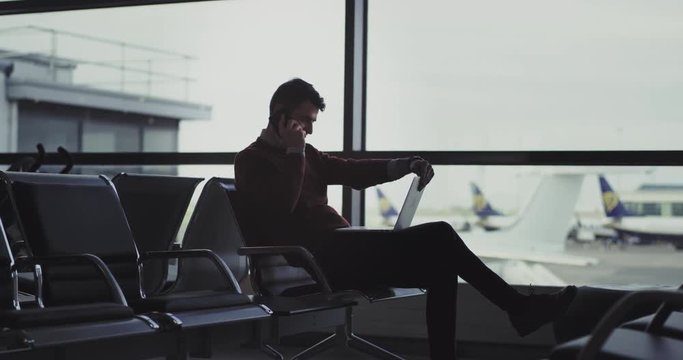 Charismatic guy in the airport sitting on the chair and speaking on his smartphone and start to work on his laptop at the same time very excited he waiting to get boarding