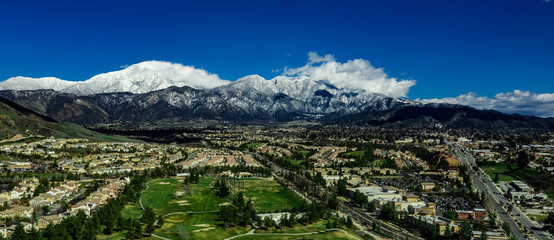 Panoramic, drone view of snow covered Mount San Gorgonio and the Little San Bernardino Mountains above Yucaipa Valley with white clouds, blue sky and green hills