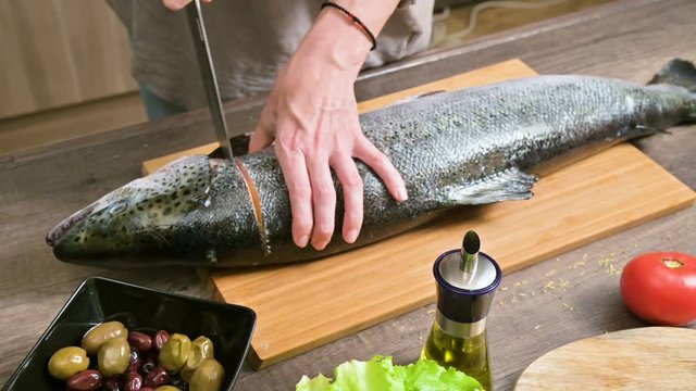 Close-up of female hands are cutting with a knife a large salmon on a wooden table of home cooking