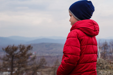 six-year-old child in a red down jacket stands in the mountains with his back to the camera