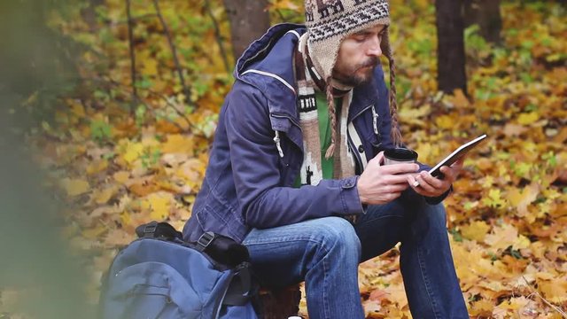 millennial bearded hipster blogger in knitted hat and scarf sitting in forest on stump, drinks hot tea or coffee and working on tablet computer, backpack near him, on background of autumn foliage 
