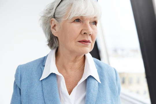 People, age, lifestyle, fashion and retirement concept. Picture of elegant fashionable sixty year old businesswoman with wrinkled face and white hair thinking over business issues, posing at window