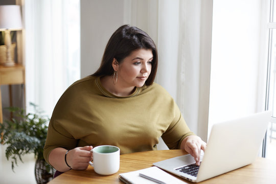 Chubby attractive young female freelancer wearing elegant sweater and round earrings working in front of open laptop, sitting in cozy home office interior, drinking coffee, browsing websites