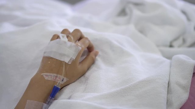 Close up IV drip solution in a patient's hand while lying on bed in hospital.