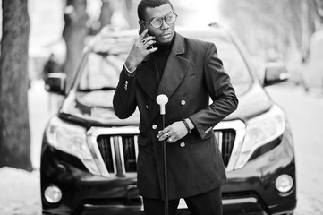 Stylish african american gentleman in elegant black jacket, holding retro walking stick as cane flask or tippling cane. Rich fashionable afro man against business suv car speaking on mobile phone.