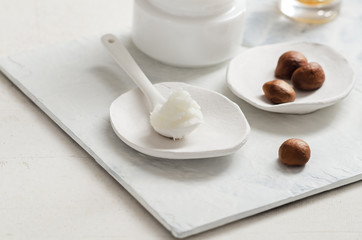 Shea butter in ceramic spoon on white background with nuts and moisturizer creme. Free text space.