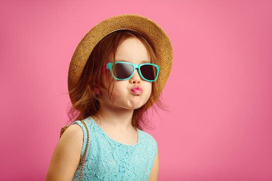 Beautiful little girl dressed in fashionable clothes, wears straw hat, sunglasses and blue dress, shows duck face, stands on pink isolated.