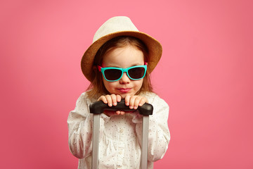 Caucasian little girl in sunglasses and straw hat, holding a suitcase, standing on isolated pink.