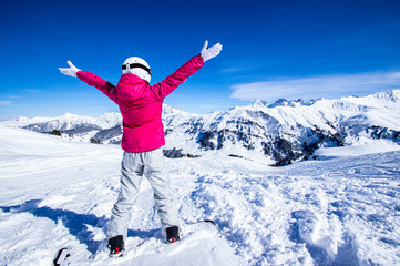 Snowy mountain view. Young happy woman snowboarder standing on the top of the mountain rising arms to the blue sky