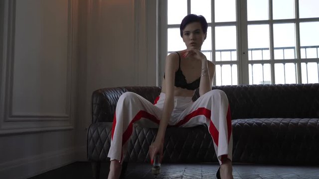 A woman with short hair, dressed in white pants with red stripes and a black bra sitting on the couch legs wide apart in her hand she has a glass of champagne, a woman posing for the camera