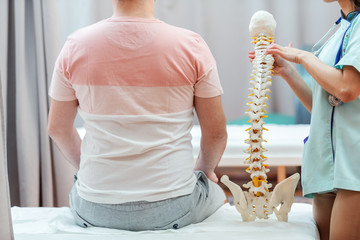 Female doctor holding spine model and pointing on vertebra while patient sitting on the hospital...