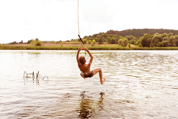 Teenage boy jumping in the river from the swinging rope on sunny summer day.