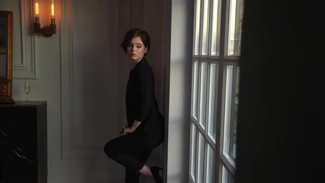 A woman with a short haircut and brown hair in a black pantsuit, with a jacket on her naked body goes leaning on the white wall and posing for the camera. Shooting in the loft with a large window