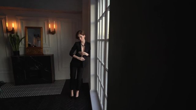 A woman with a short haircut in a black pantsuit, with a jacket on her naked body goes leaning on a white wall and posing for the camera. Shooting in a beautiful room with a large window