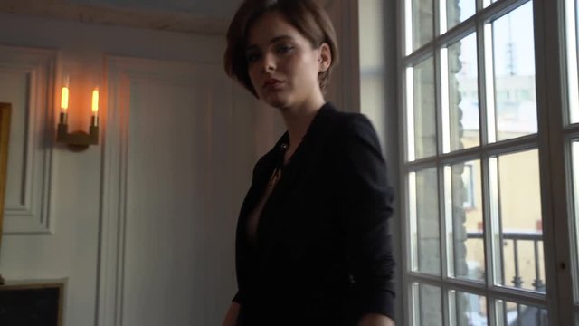 A woman with a short haircut, dressed in a black jacket on a naked body goes to the camera, holds a glass of champagne in her hands, stops and drinks champagne, then goes back to the camera and stops 