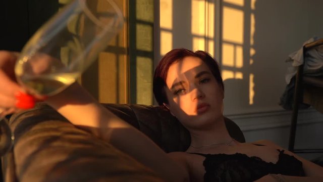 A woman with short black hair, wearing white pants and a black bra lies on a black couch and looks out the big window at the sunset. On the face of the fall sunshine, a girl holds a champagne glass