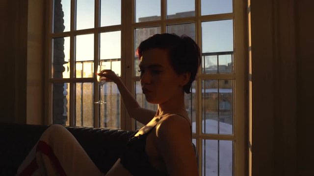 A woman with a short haircut and dark hair, dressed in a black bra lies on a black sofa at the big window in her hands holding a glass of champagne, drinking it, dripping champagne on the body