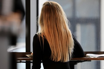 młoda kobieta blondynka siedzi tyłem w kawiarni miło spędza czas pije kawę czyta gazetę - obrazy, fototapety, plakaty